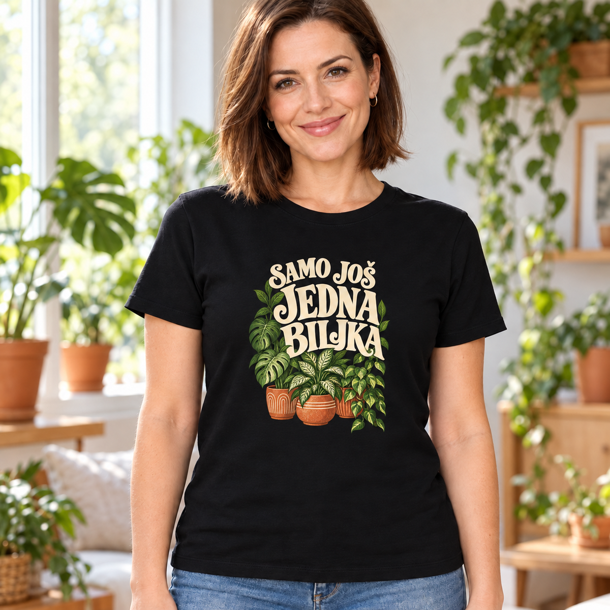 Žena u crnoj majici sa krem natpisom „Samo još jedna biljka“ i zelenim sobnim biljkama u tri terakota saksije, u svetlom enterijeru.